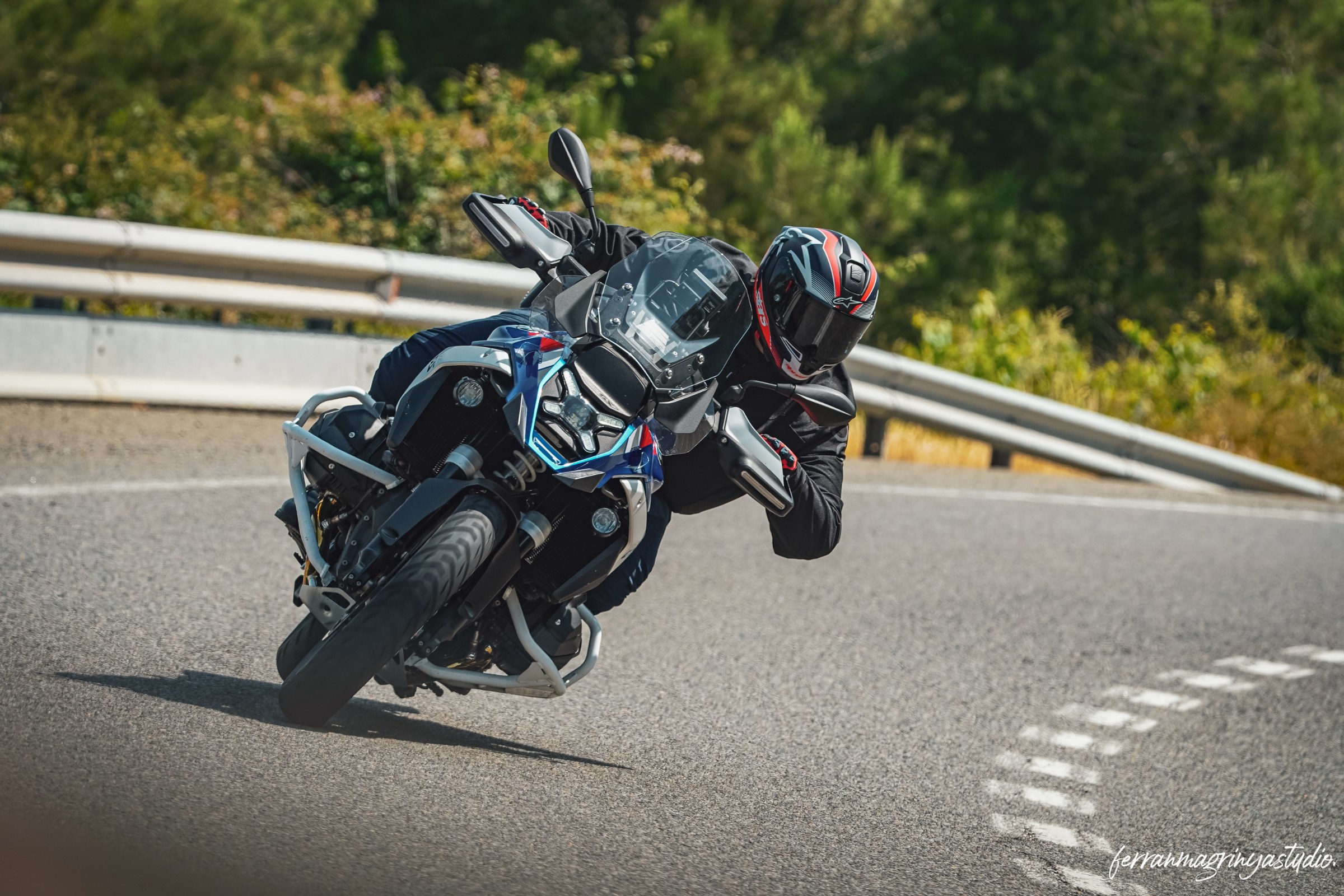 Una moto azul y su motorista inclinándose en una curva, con vegetación al fondo.
